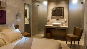Quarto de hotel com uma cama grande, área de estar com uma poltrona, banheiro com pia e espelho, e um objeto decorativo na parede.