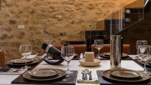 Uma mesa de jantar posta com pratos, copos, talheres e uma garrafa de vinho, com uma parede texturizada e um corrimão de vidro ao fundo.