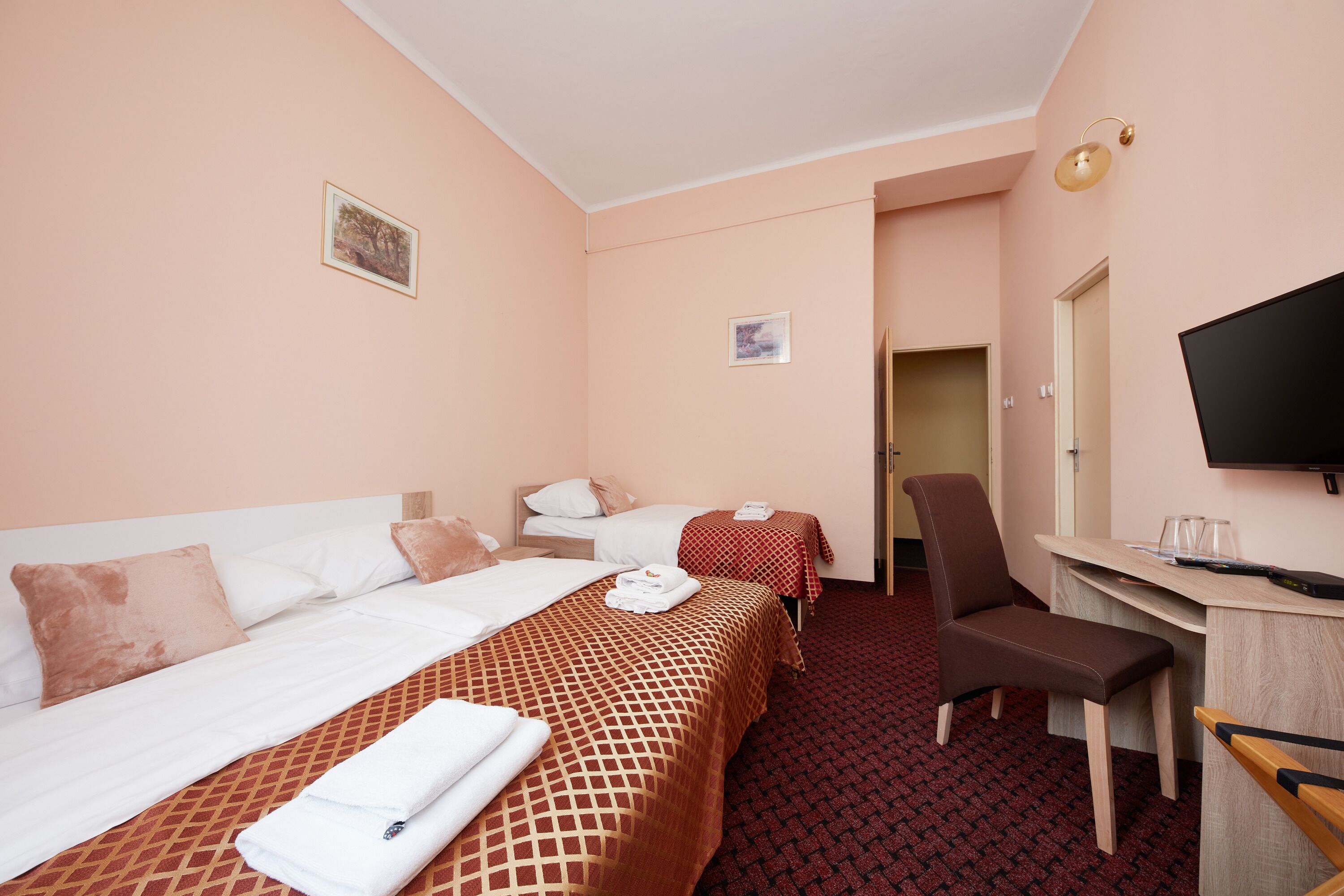 A hotel room with two beds, a desk, a chair, a television, and a door leading to another room.