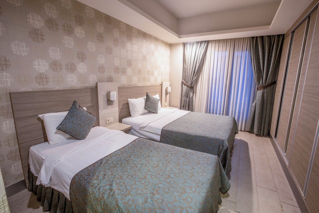 A hotel room with two beds, a large window with curtains, and a patterned wallpaper.