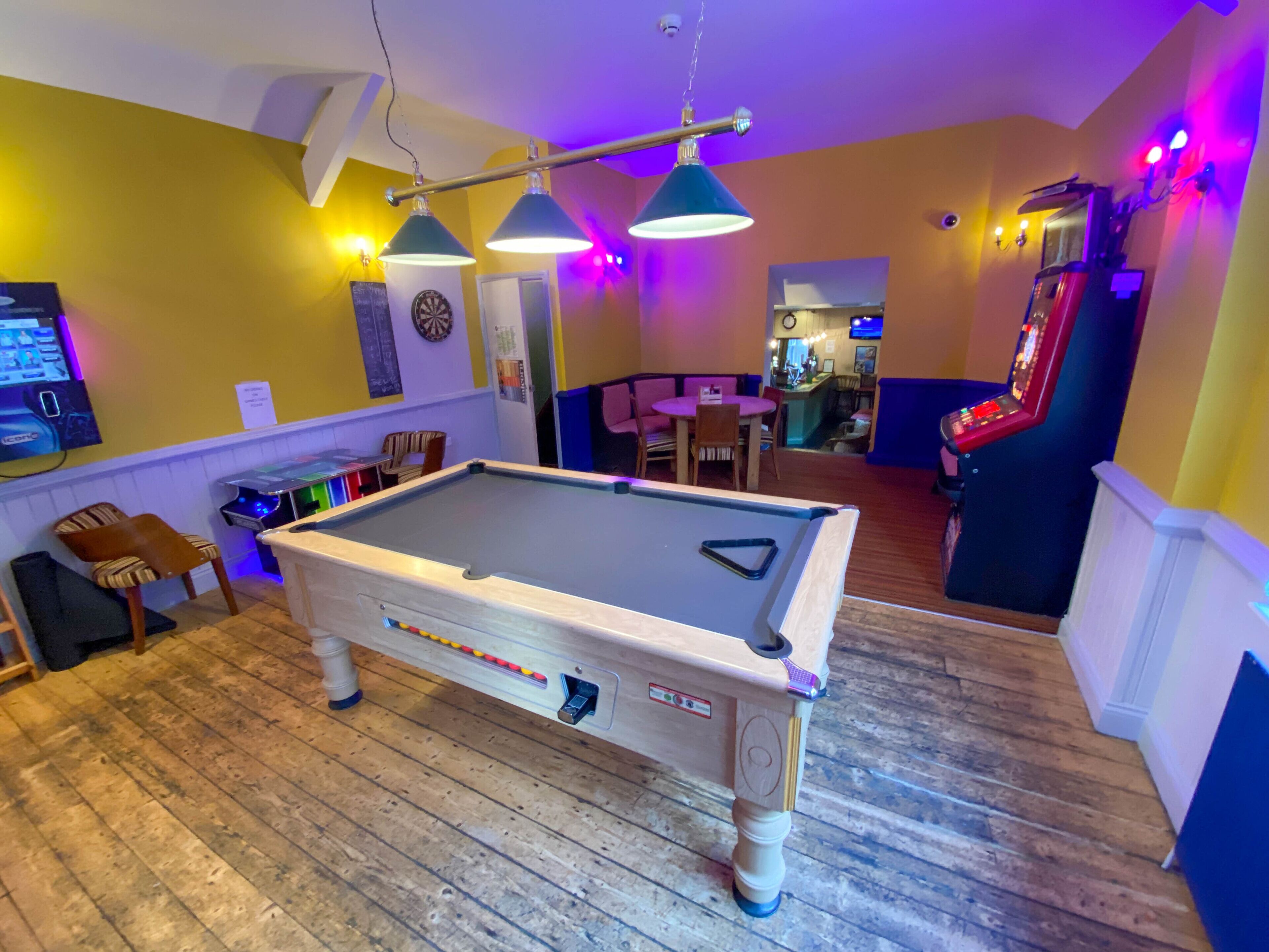 Une table de billard se trouve dans une pièce dont le sol est en bois, les murs sont peints en jaune et un jeu de fléchettes est accroché au mur. Au fond, on aperçoit un bar avec un comptoir et des tabourets.