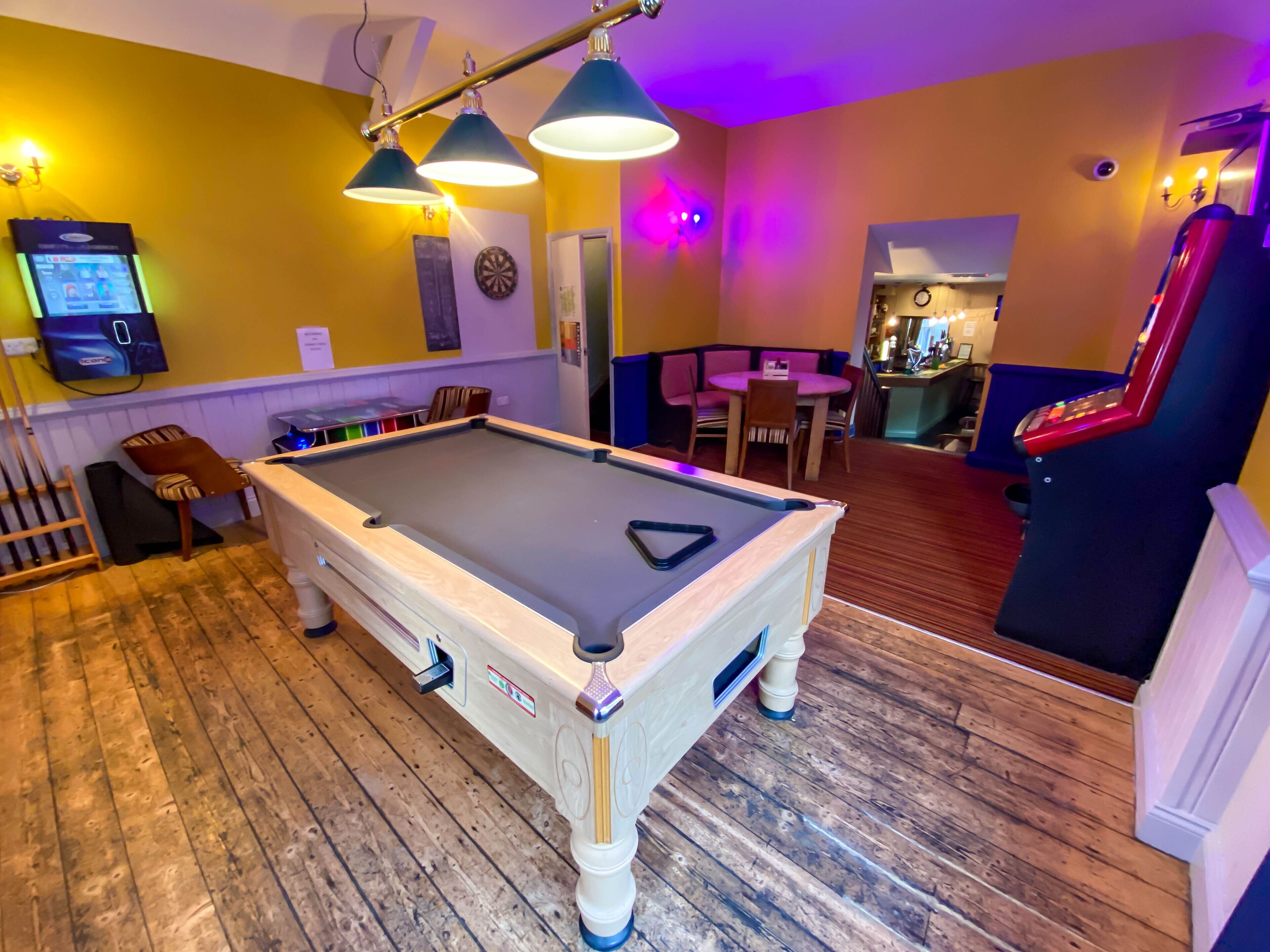 A pool table in a room with wooden flooring, a dartboard, and a billiards table.
