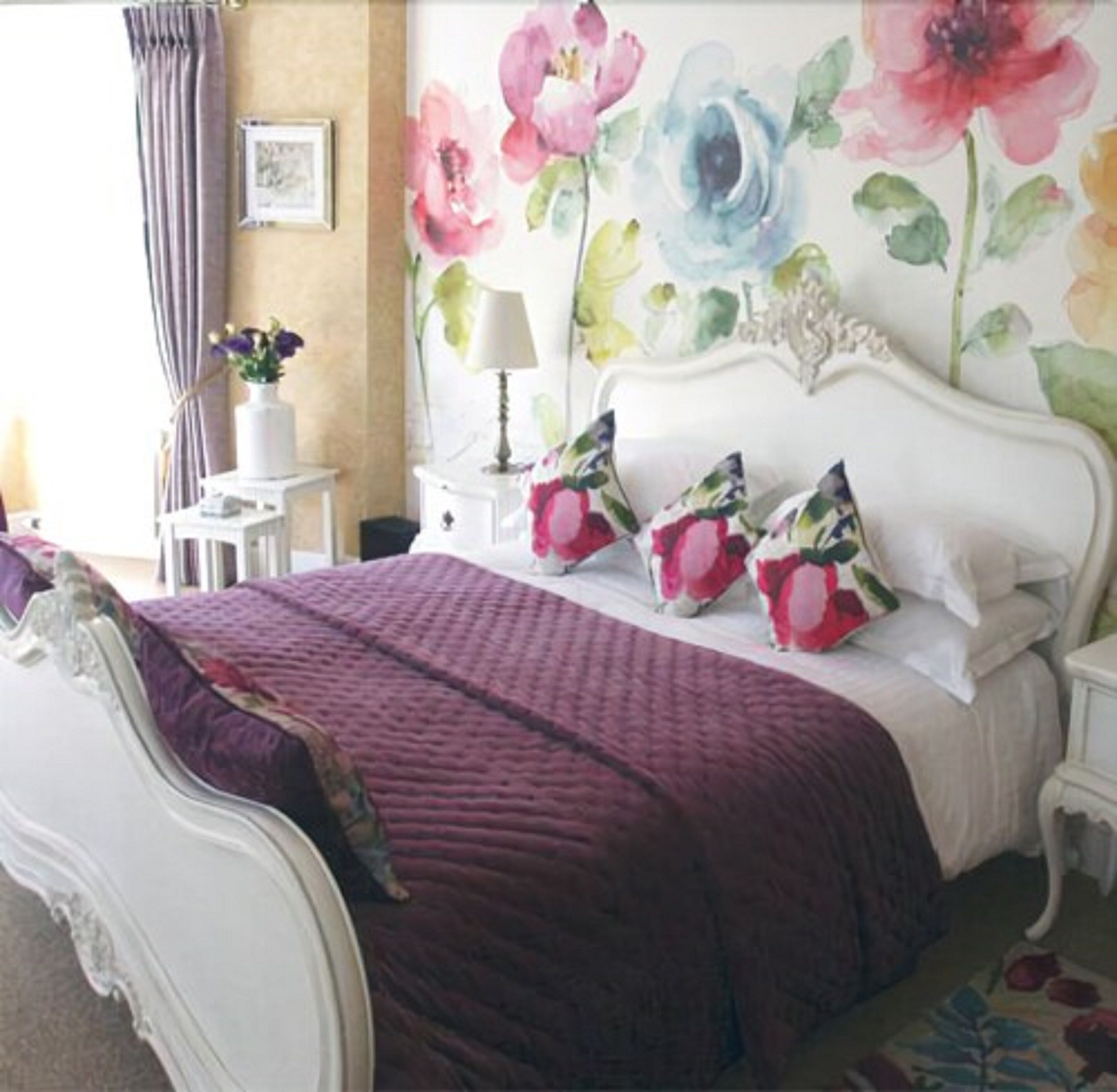 Un dormitorio con cama blanca, ropa de cama morada y decoración floral en las paredes.