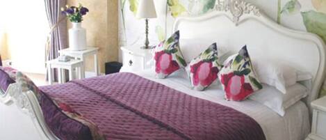 A bedroom with a white bed, purple bedding, and floral wall art.