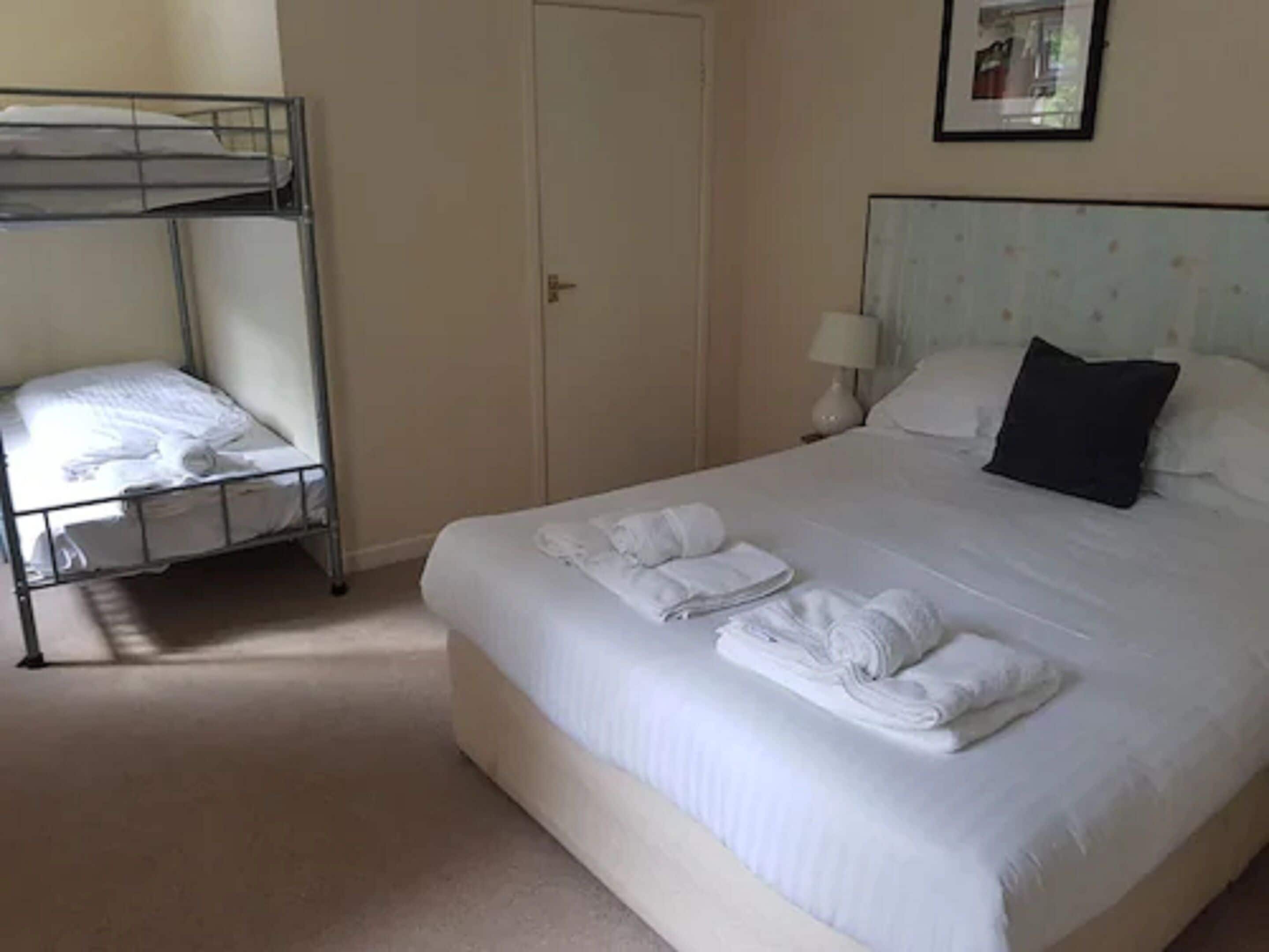 A hotel room with a bunk bed, a large bed with neatly folded towels, a nightstand with a lamp, and a framed picture on the wall.