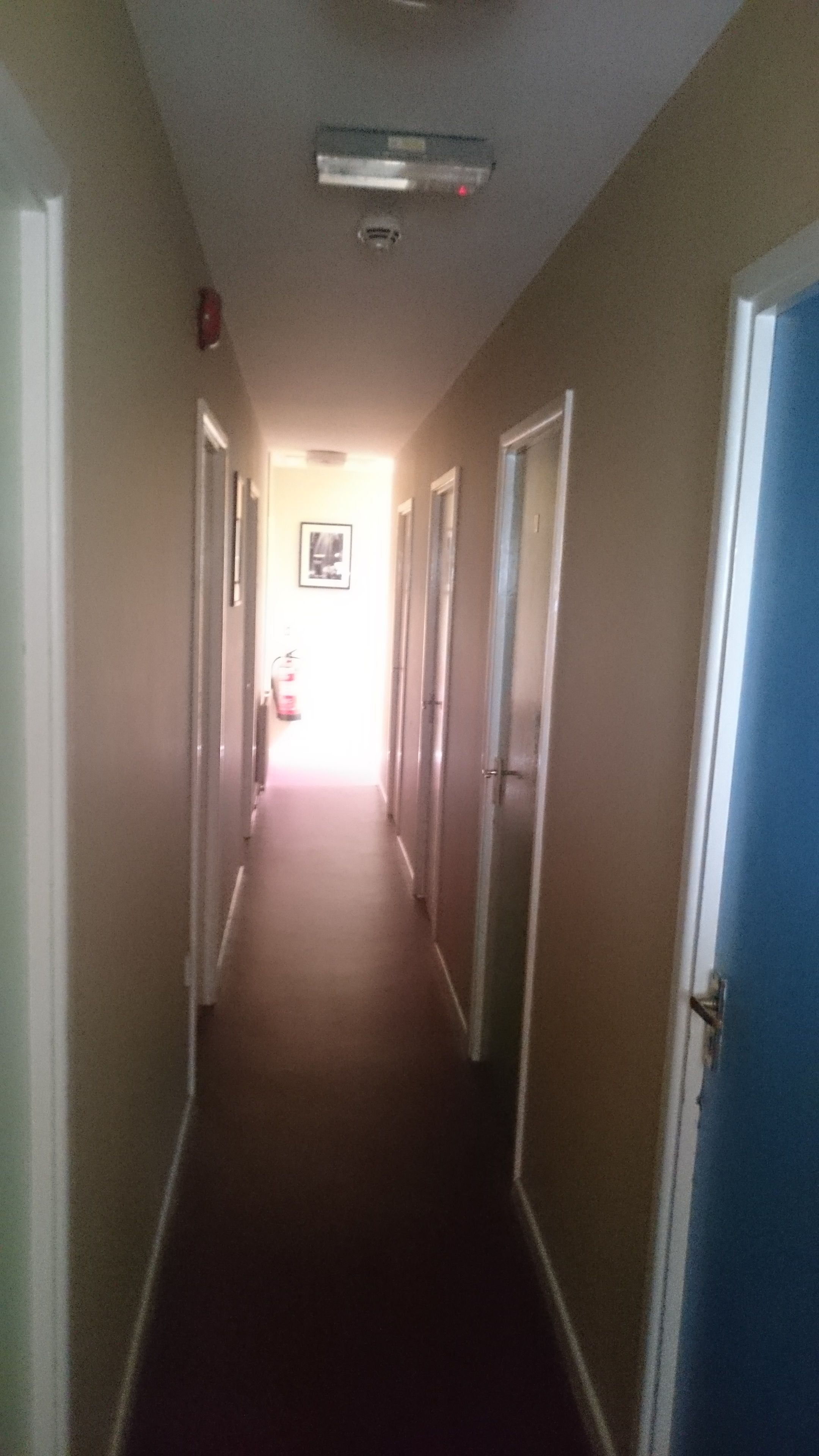 A narrow hallway with doors on both sides, a ceiling light, and a door at the end leading to a brighter area.