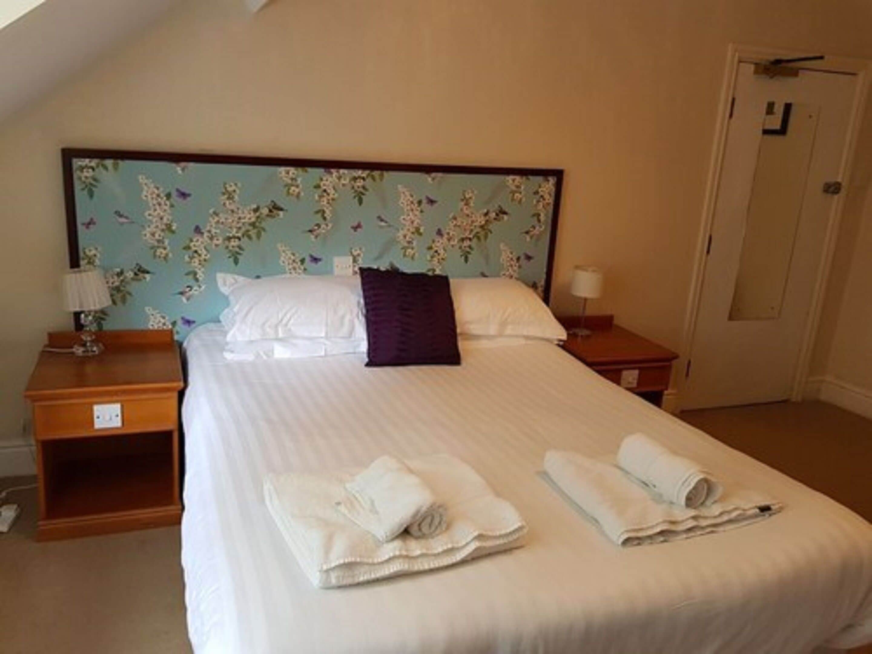 A neatly made bed with white linens, a floral headboard, and two rolled towels on the bed.