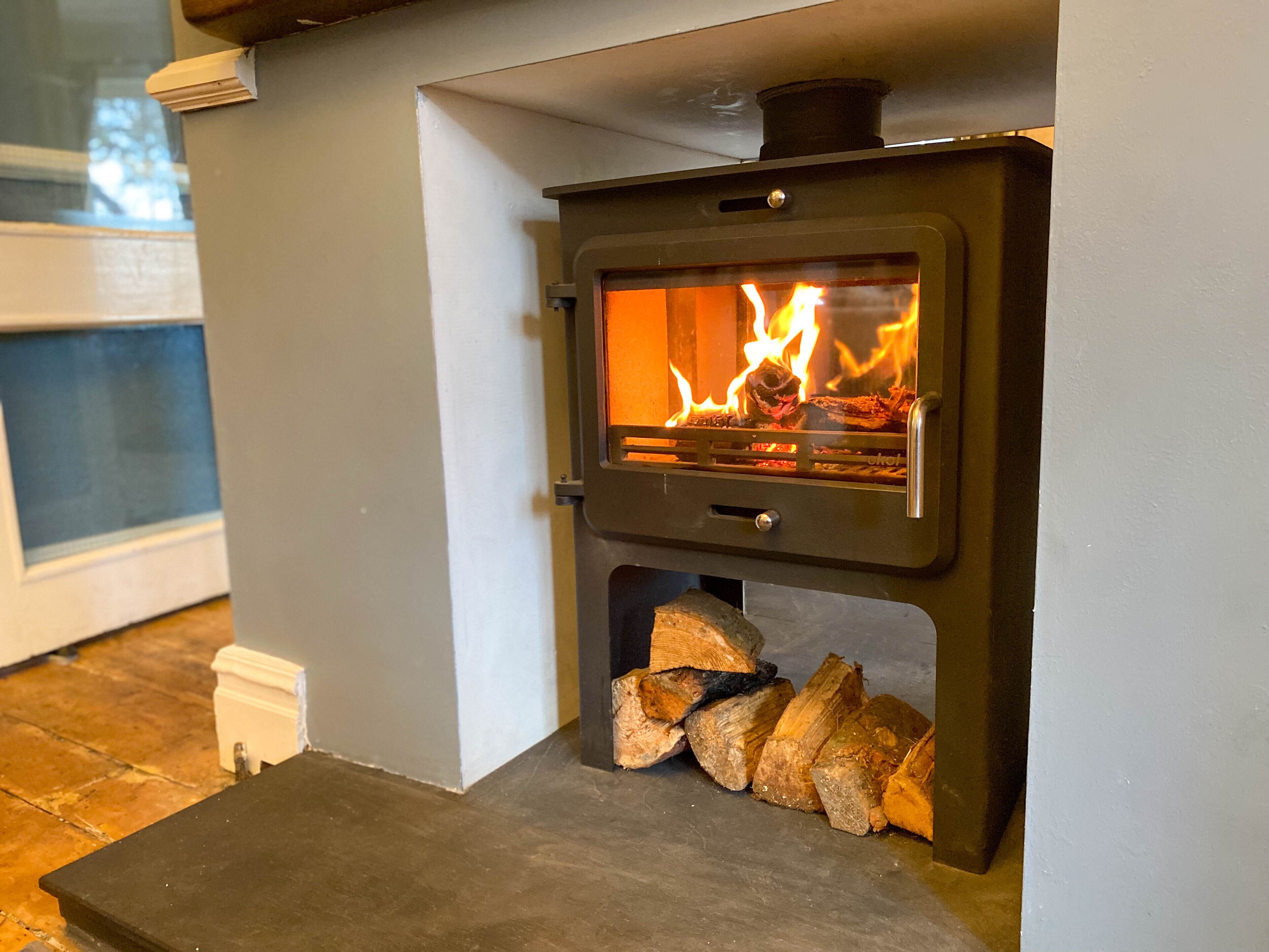 A wood-burning stove with visible flames, a storage compartment below, and a stack of firewood.