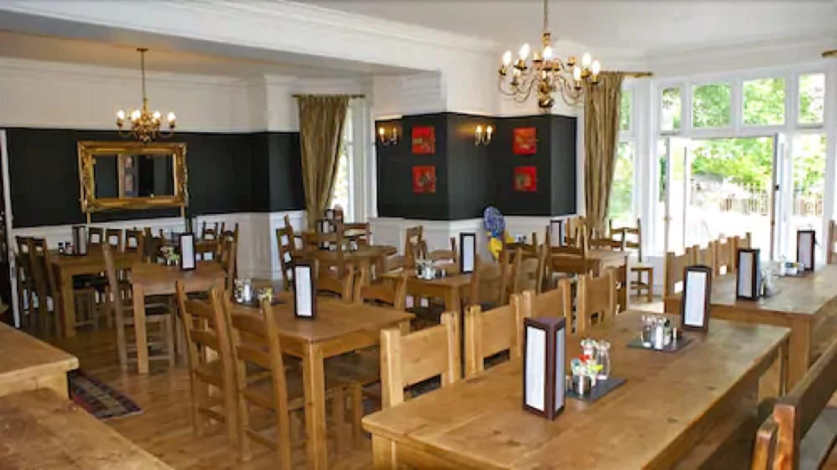 A spacious dining area with wooden tables and chairs, large windows, and a chandelier.