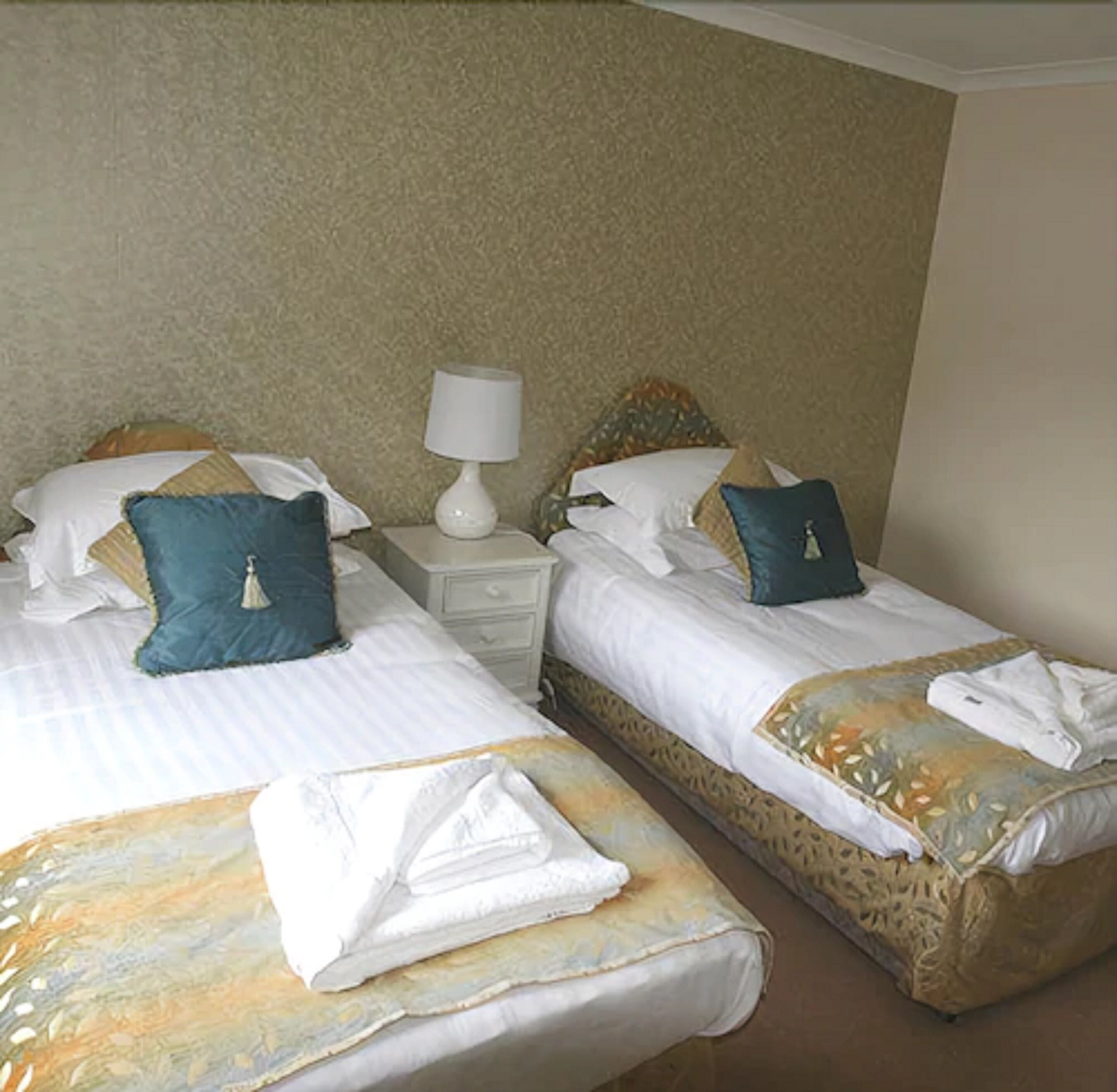 Two single beds with white linens, blue pillows, and a white bedside table with a lamp.