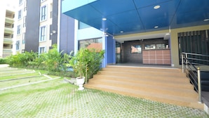 A modern building entrance with a blue awning, a staircase leading to a ground floor, and a well-maintained garden area with potted plants. - Kwanchanok Ville (Hua Hin)