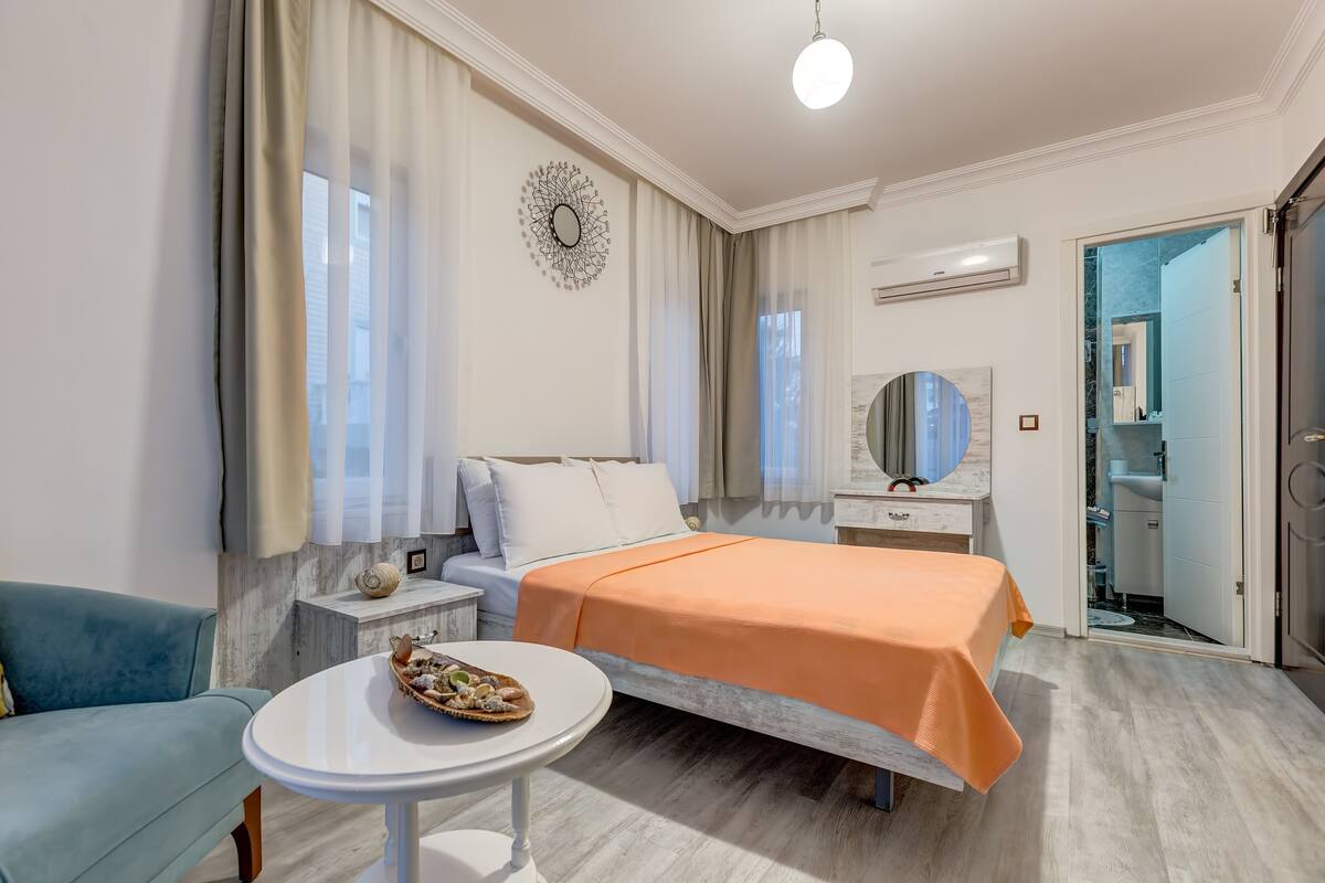 A bedroom with a bed, bedside tables, a round table, a blue armchair, and a bathroom visible through an open door.