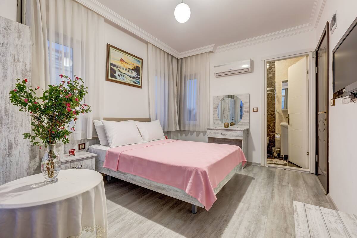 A bedroom with a bed, bedside table, and a mirror. There is a window with curtains, a painting on the wall, and an air conditioning unit.