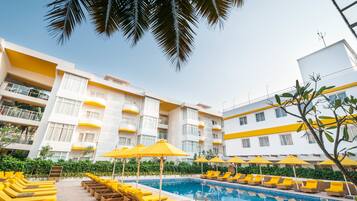 Zona de piscina de hotel con tumbonas amarillas y sombrillas, rodeada por un edificio blanco con balcones.