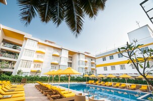 A hotel pool area with yellow sun loungers and umbrellas, surrounded by a white building with balconies.