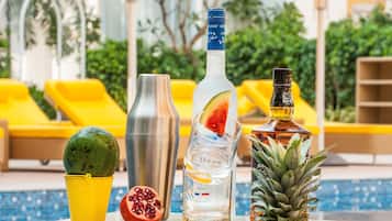 A poolside setting with a cocktail glass, a bottle, a pomegranate, and a pineapple on a glass table.