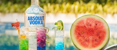 Eine Flasche Absolut Wodka, drei farbenfrohe Cocktails und ein halbiertes Wassermelone am Pool.