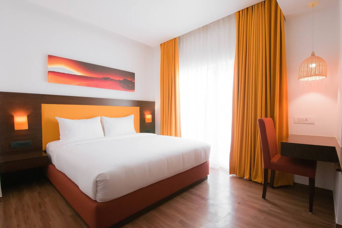Habitación de hotel con cama, escritorio y silla. Hay un cuadro en la pared y una ventana con cortinas.