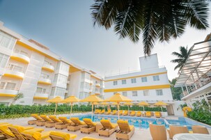 A pool area with sun loungers and umbrellas, surrounded by a building with balconies and a clear sky.