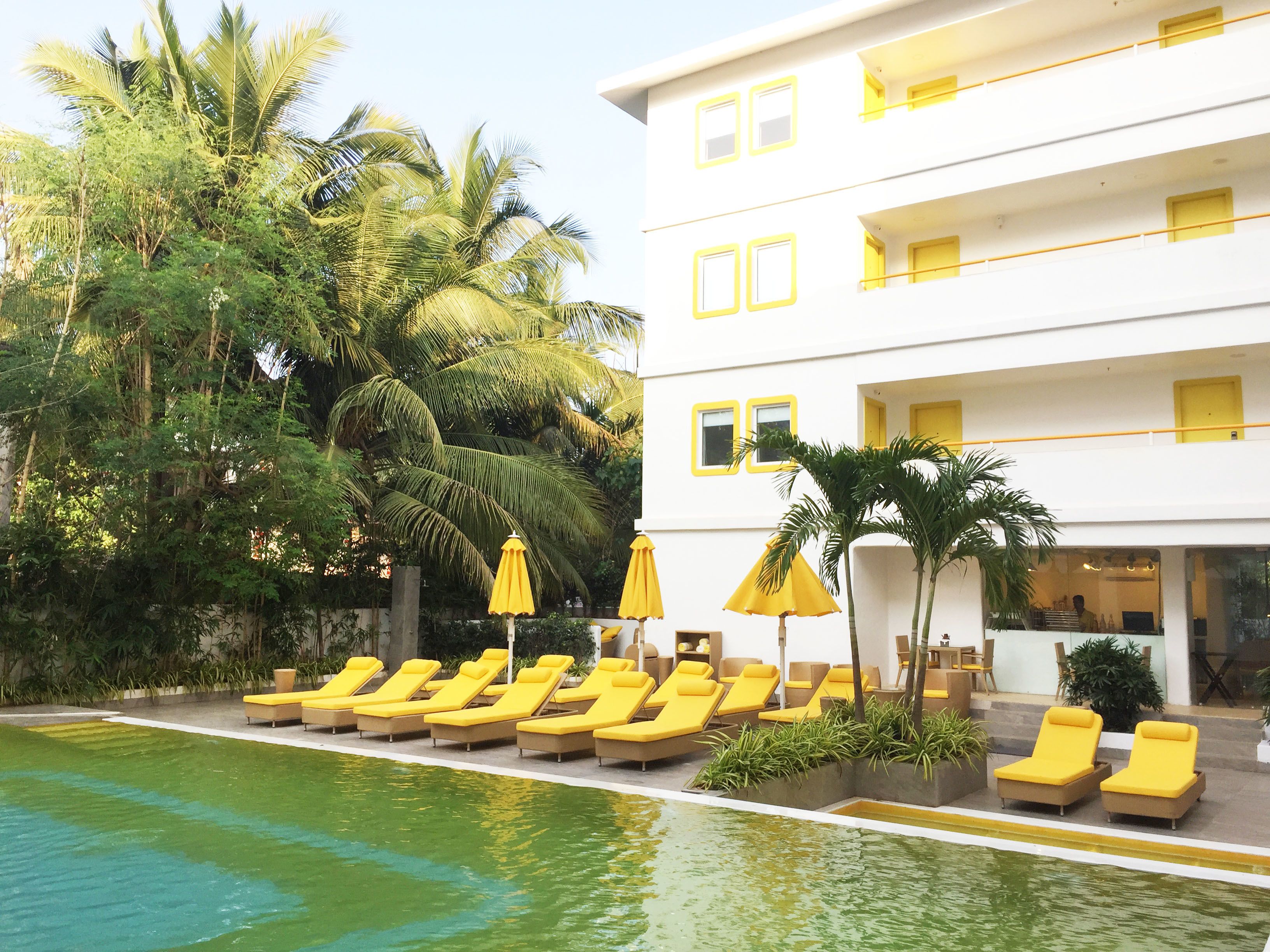A poolside area with sun loungers and umbrellas, surrounded by palm trees and a white building with yellow accents.