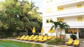 A poolside area with sun loungers and umbrellas, surrounded by palm trees and a white building with yellow accents.