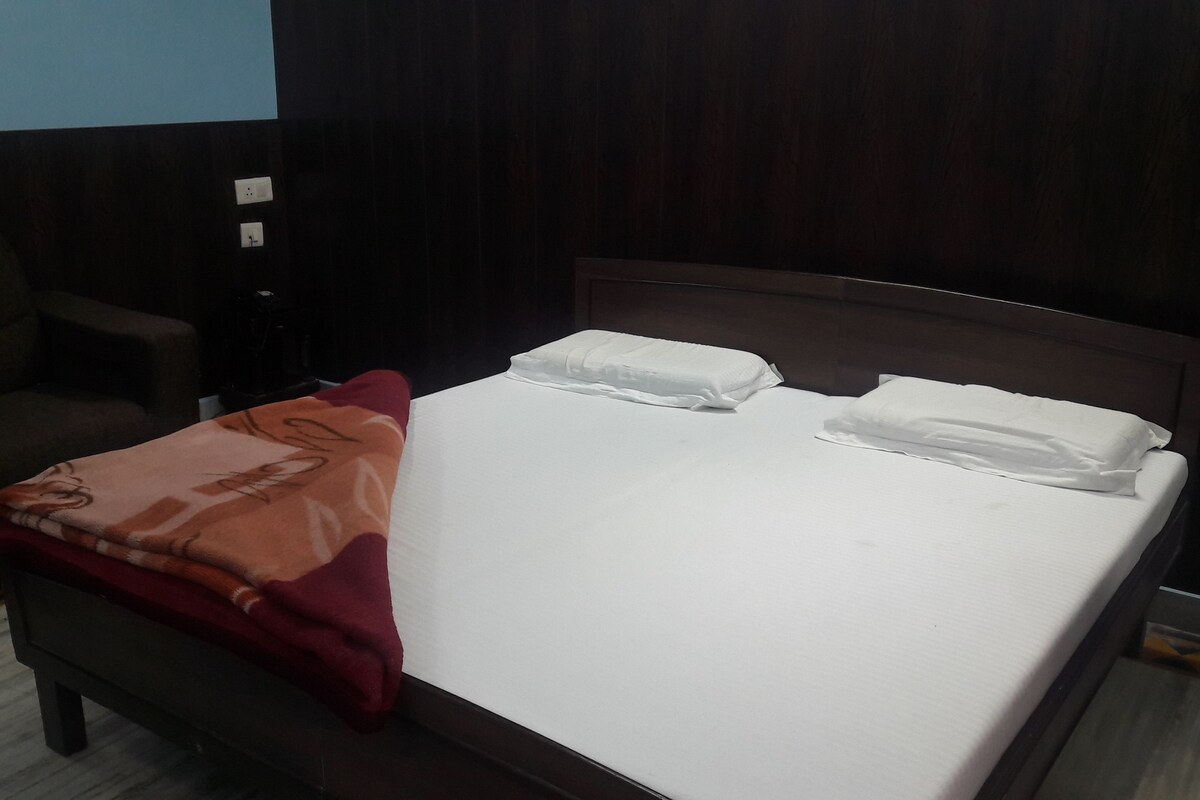 A neatly made bed with white linens and two pillows, a red blanket, and a wooden headboard in a room with dark walls and a carpeted floor.