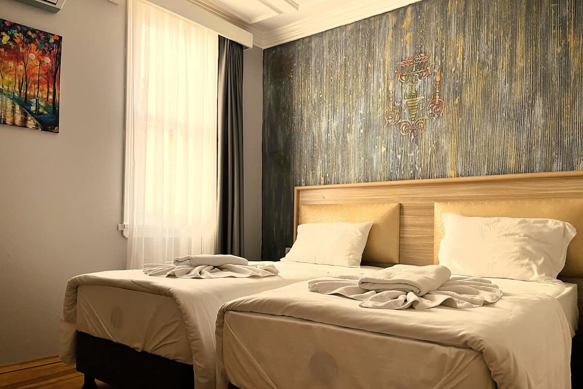 A hotel room with two beds, a large window with sheer curtains, a textured wall, and a painting on the wall.