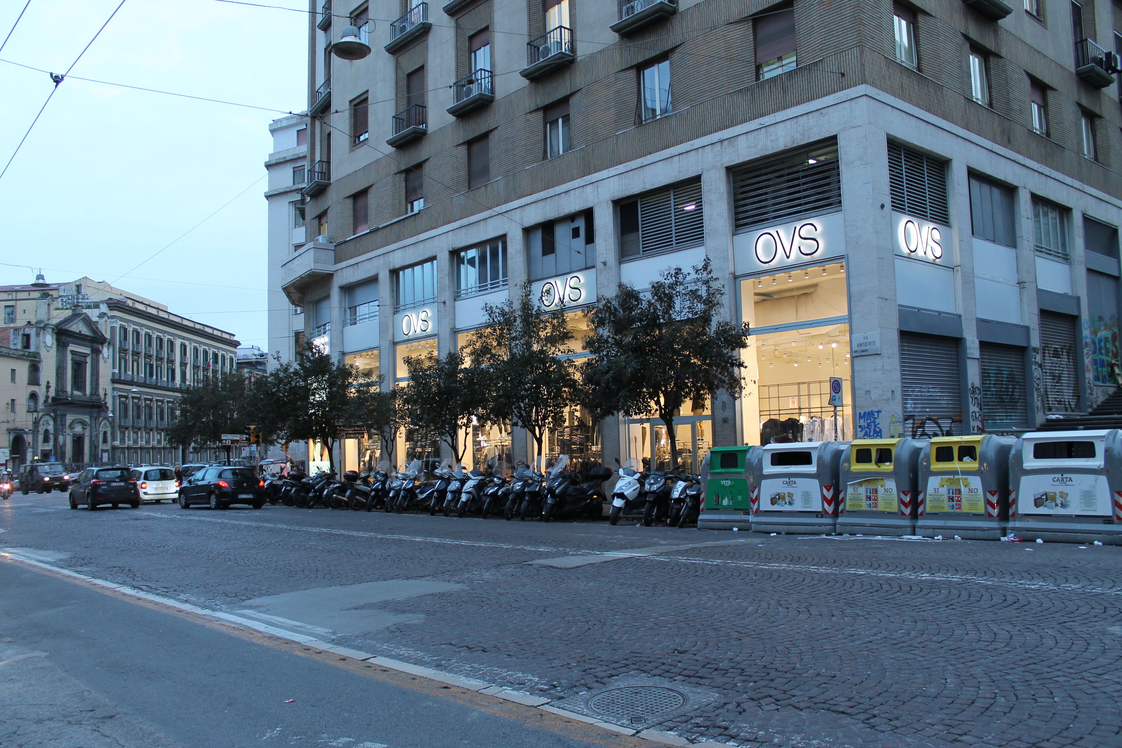 A building with multiple OVS store fronts, parked motorcycles, and a cobblestone street.