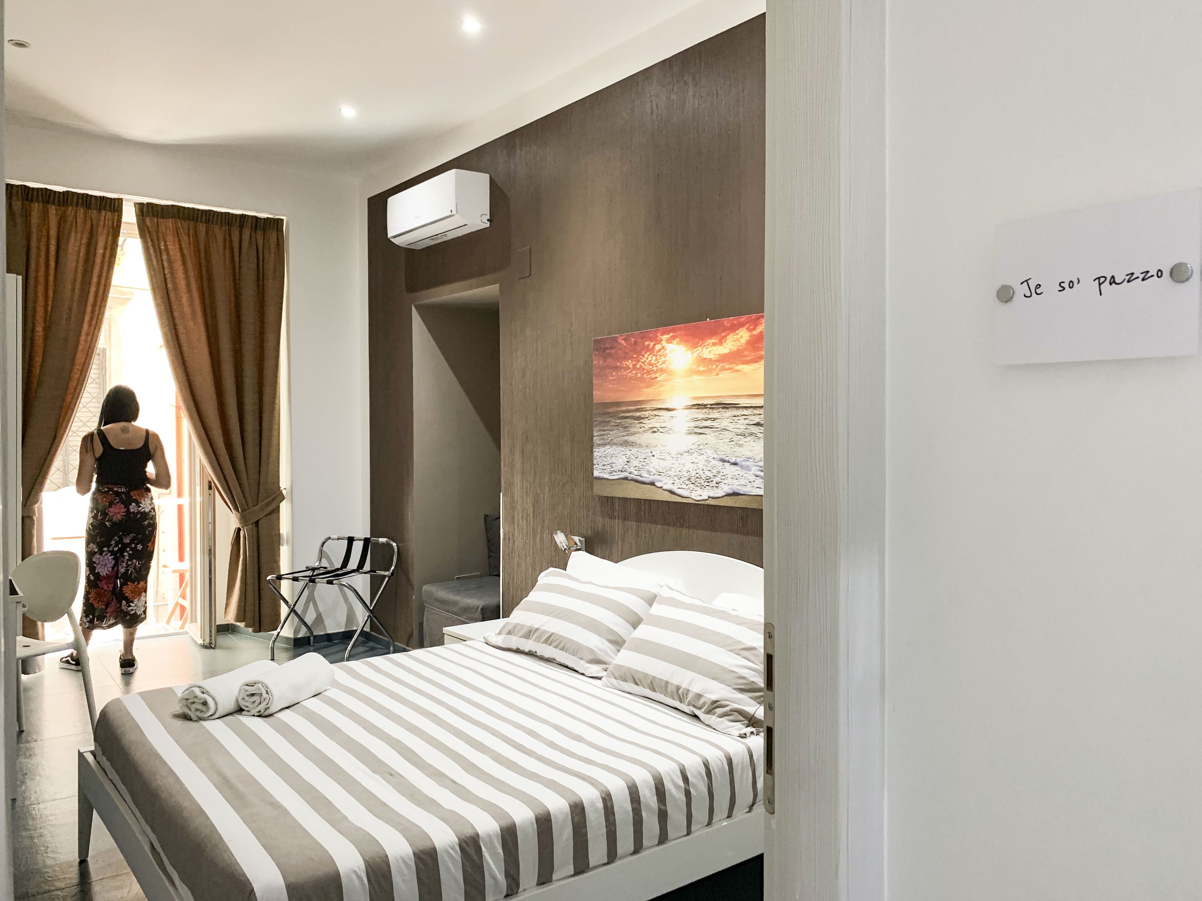 A bedroom with a bed, a chair, an air conditioning unit, and a painting of a sunset over the ocean.