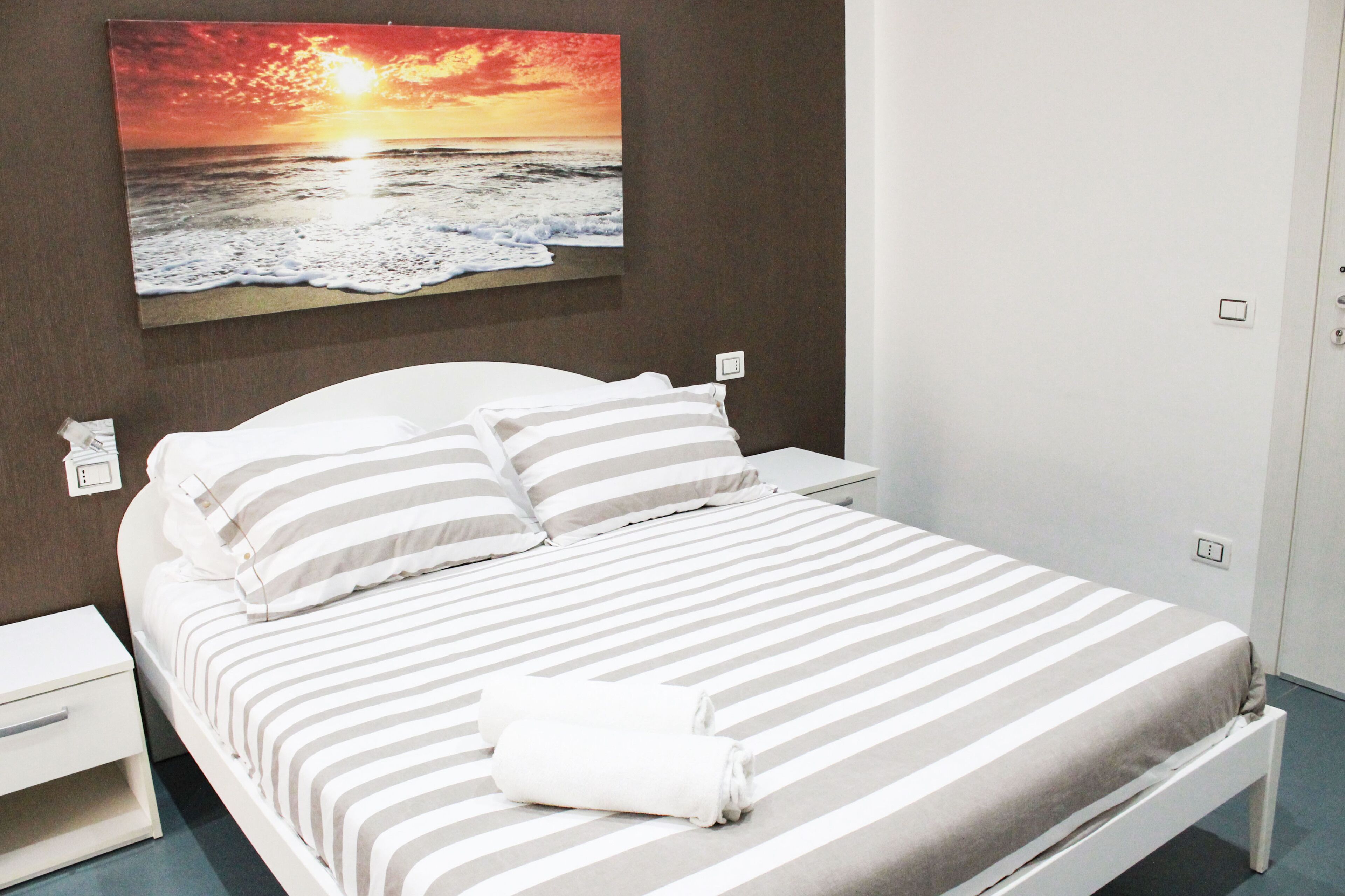 Una cama bien tendida con ropa de cama a rayas, una mesita de noche blanca y un cuadro de un atardecer sobre el océano en la pared.