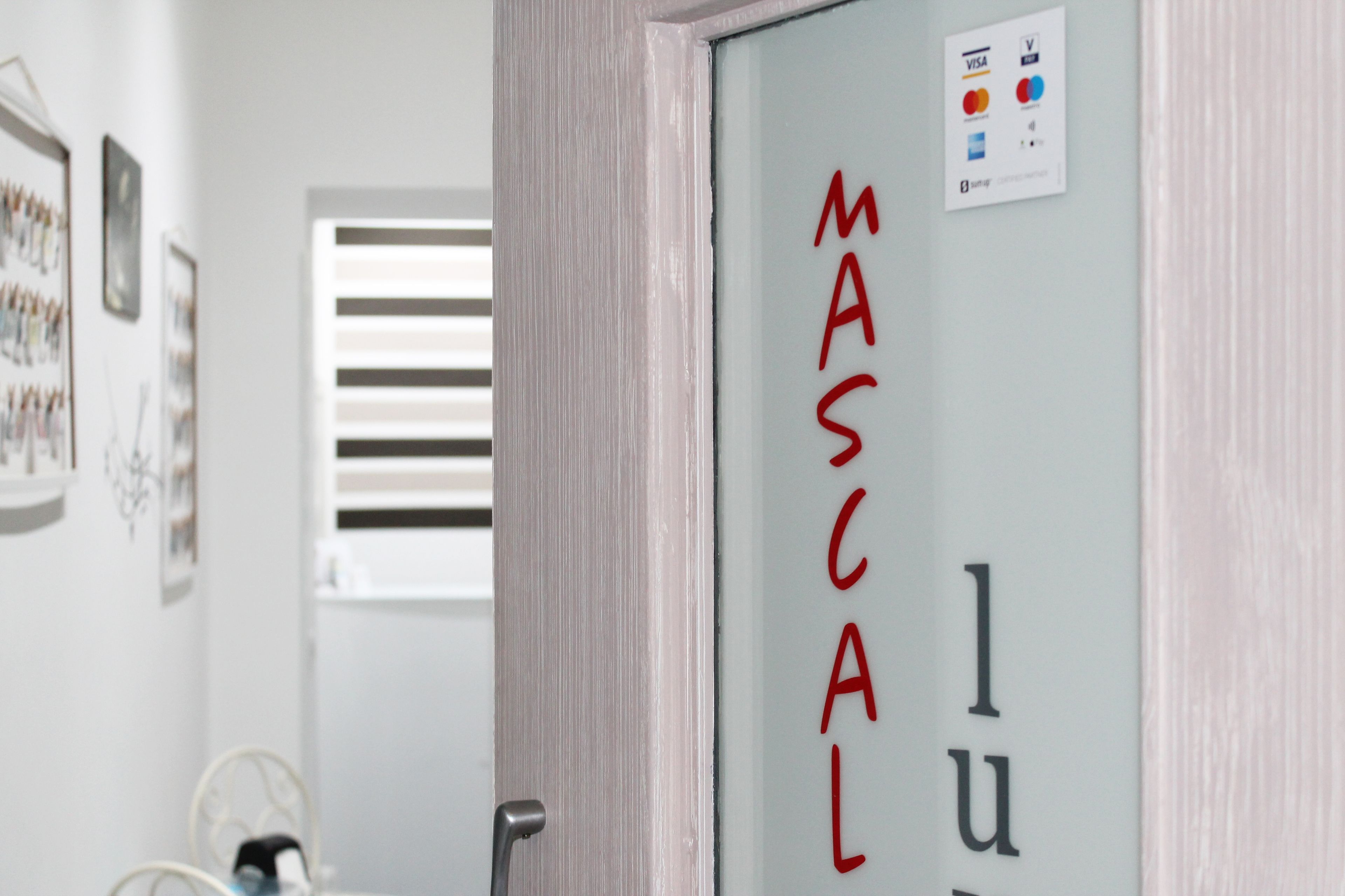 Une porte sur laquelle on peut apercevoir le mot « MASCAL », une fenêtre avec des stores horizontaux et, au fond, une chaise en métal.