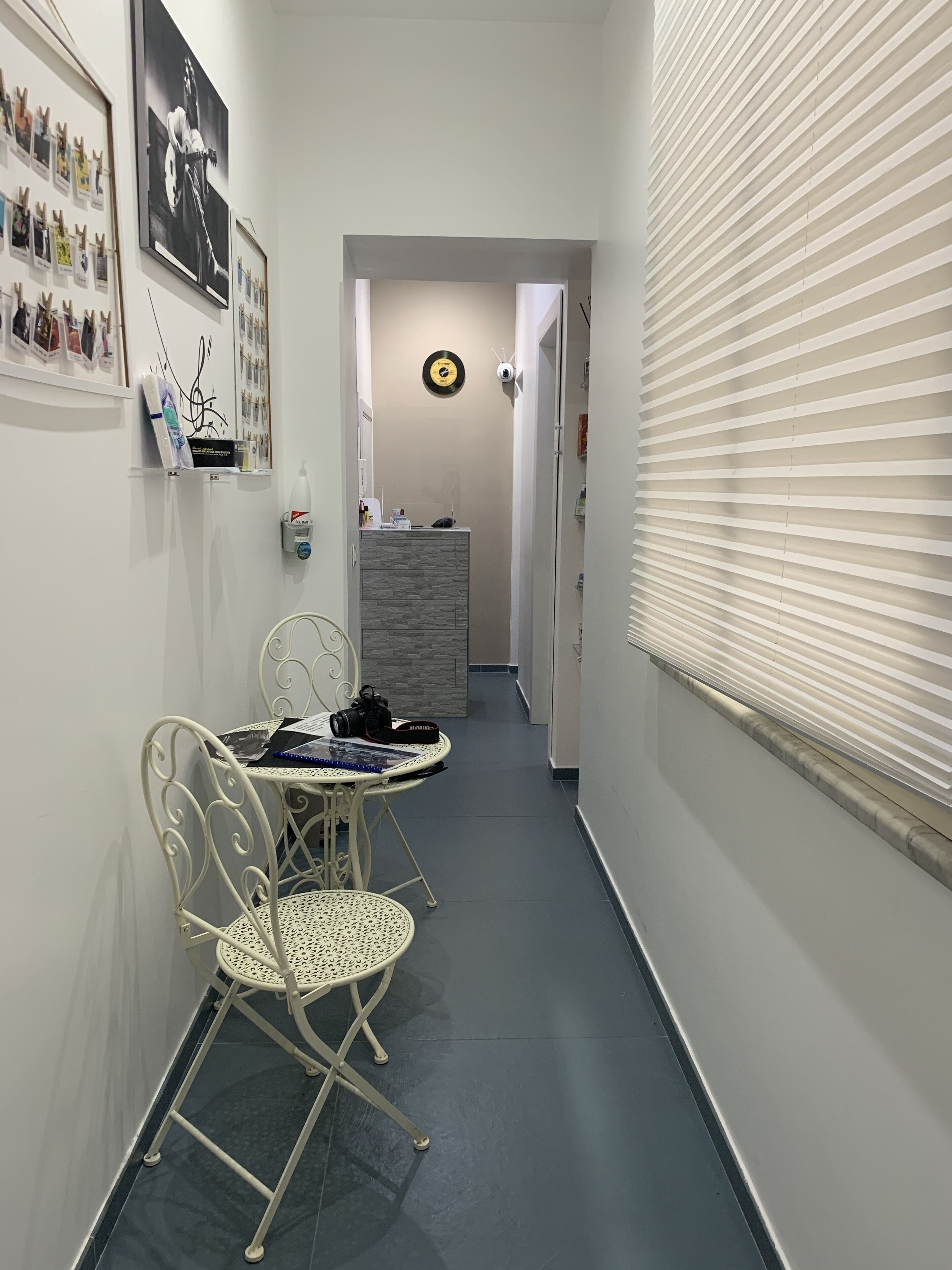 A narrow hallway with a dining area featuring a white metal table and chairs, a small cabinet with a clock, and a window with blinds.