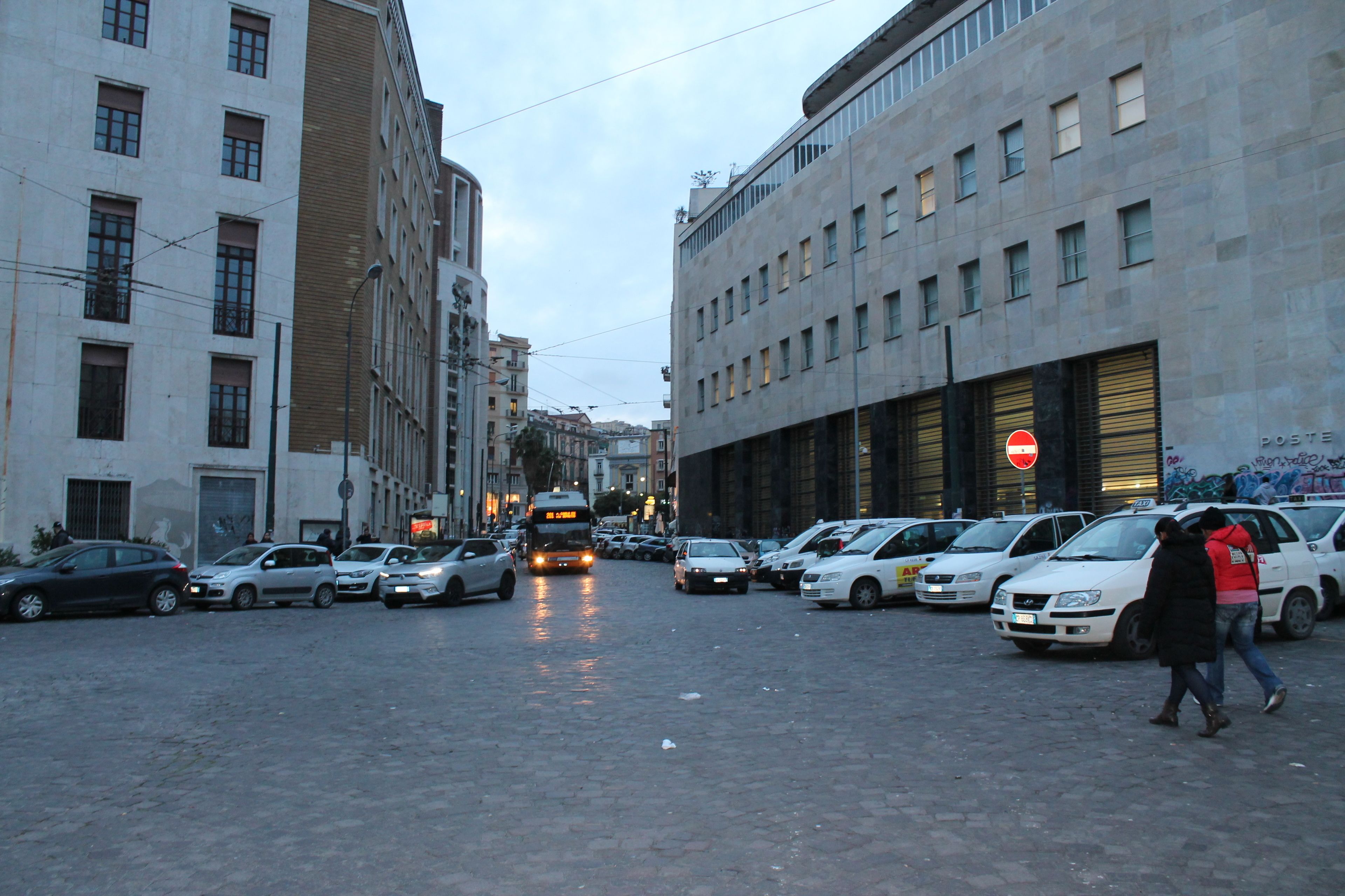 A city street with parked cars and a bus. Buildings line the street, and there are people walking.