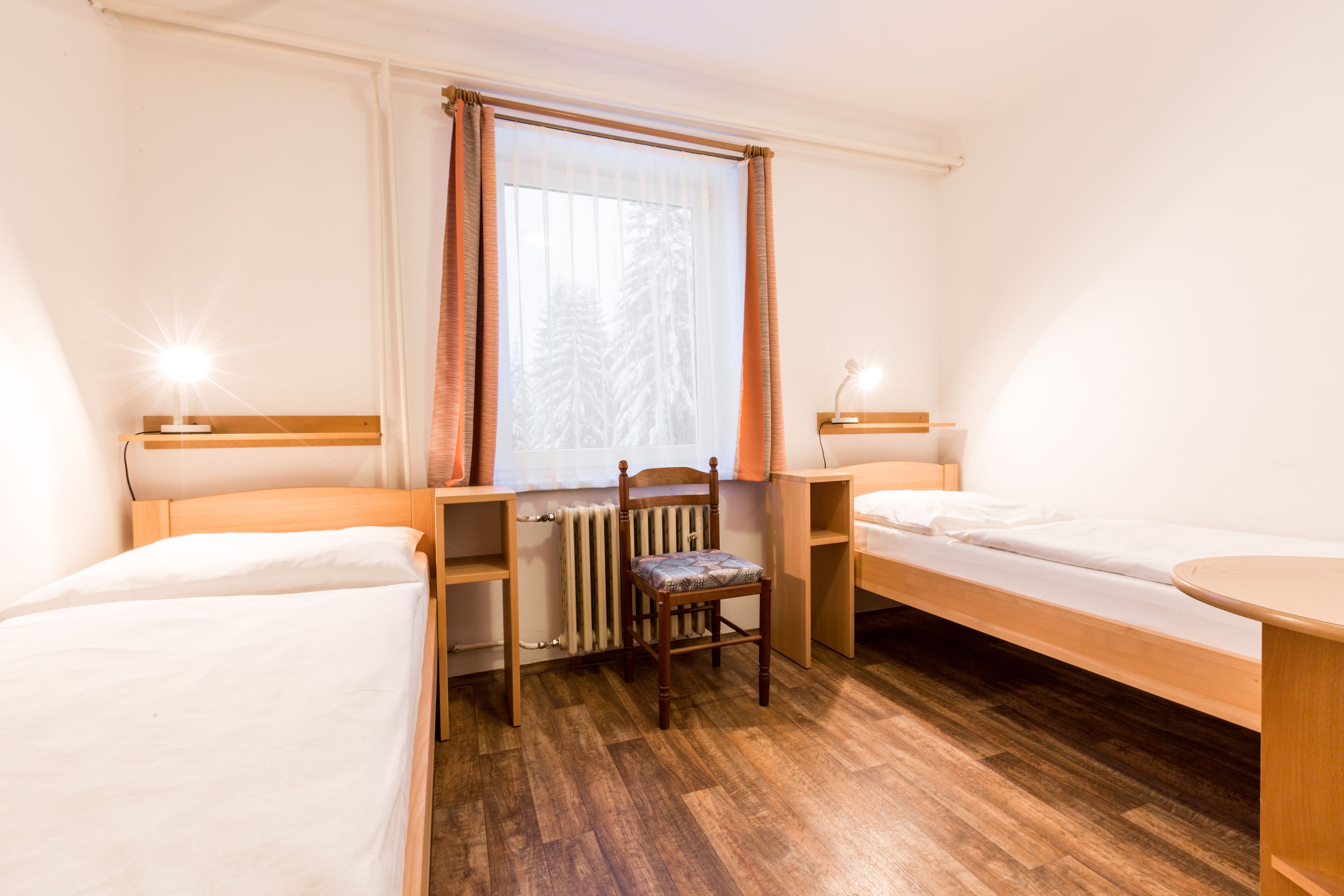 Una habitación con dos camas individuales, una silla de madera, un radiador y una ventana con cortinas.