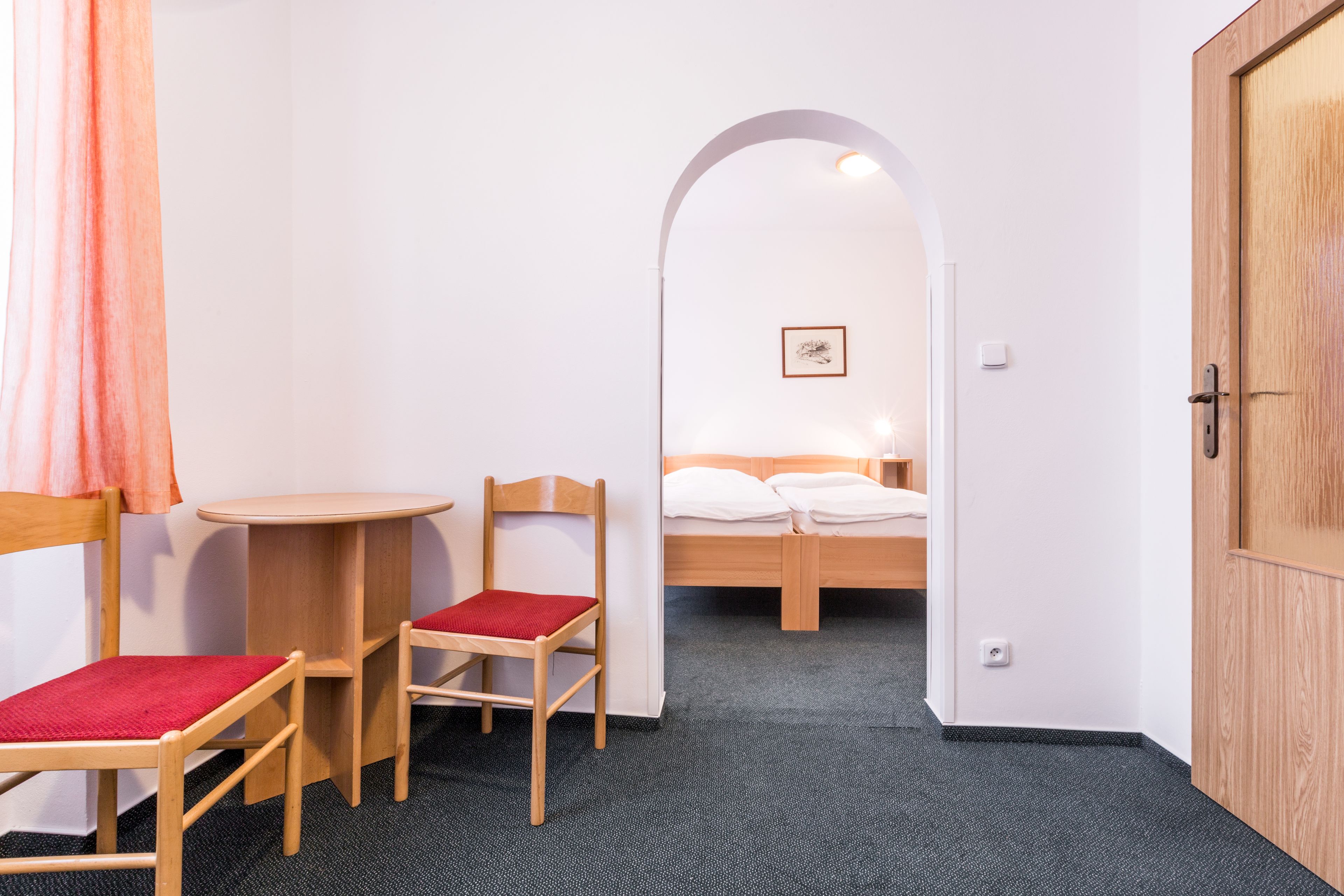 Une petite pièce comprenant un lit, deux chaises en bois avec des coussins rouges, une petite table ronde et une porte blanche.