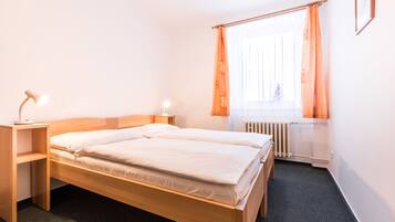 A neatly made bed with a wooden headboard, a bedside table with a lamp, a window with orange curtains, and a radiator.