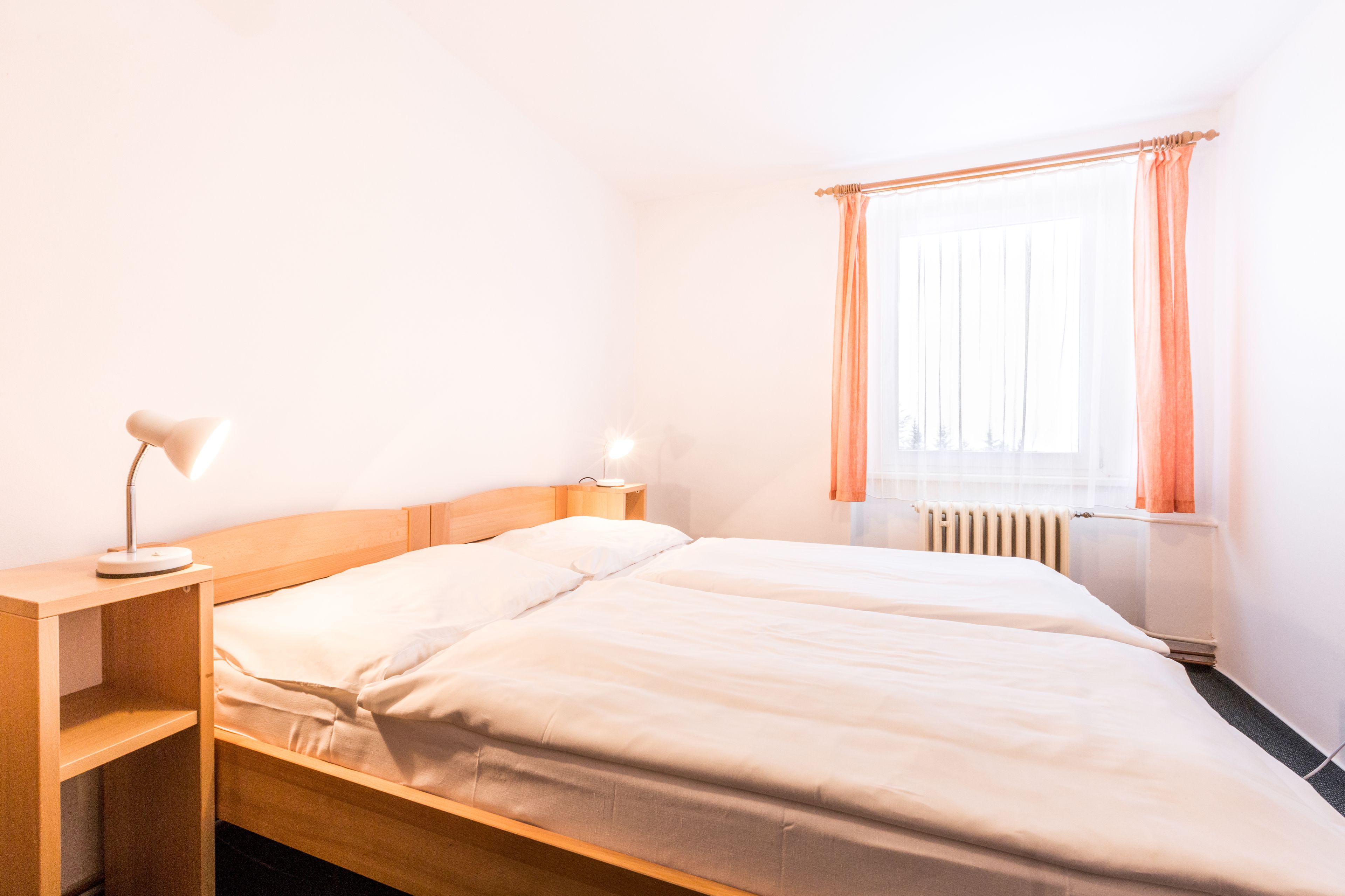 A hotel room with two beds, a nightstand, a lamp, a window with curtains, and a radiator.