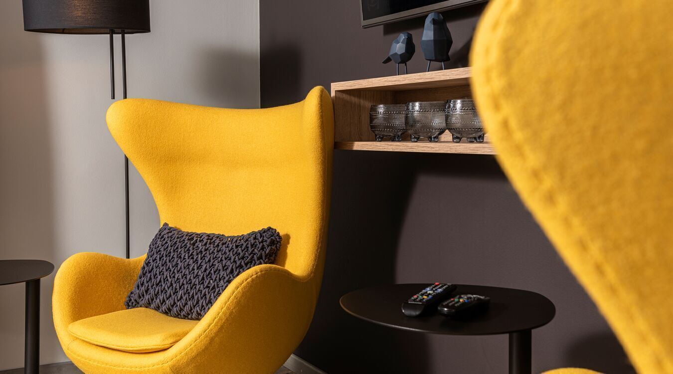 A yellow armchair with a textured cushion, a small round table with two remote controls, and a shelf with decorative items.