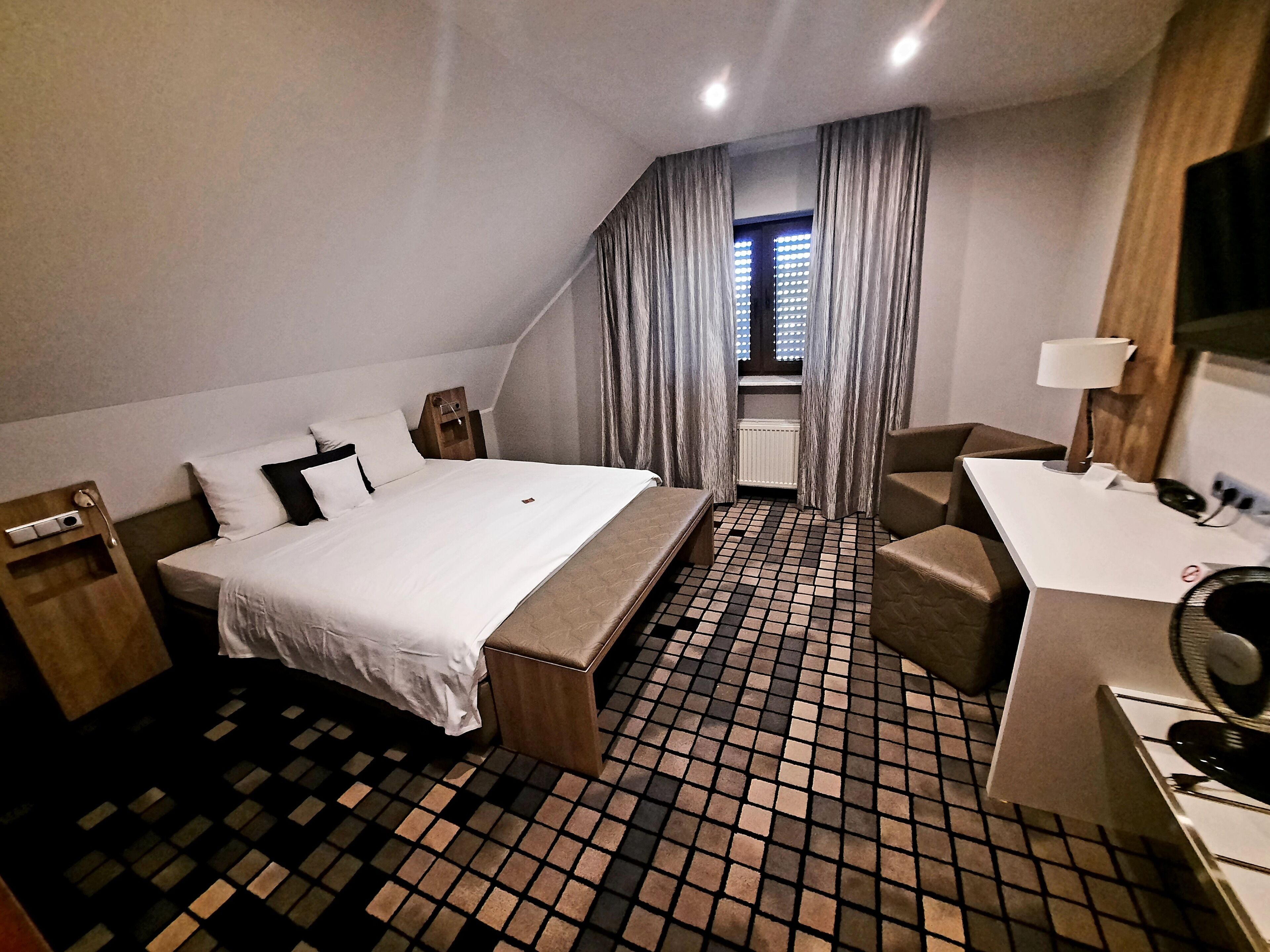 Una habitación de hotel con cama, escritorio, silla, lámpara y ventana con cortinas.