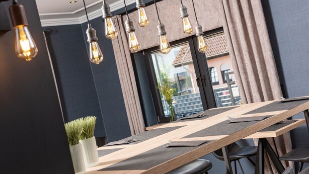 A modern dining area with a long wooden table, multiple chairs, and hanging light bulbs. - Hotel Bei Den Tongruben (Neuberg)