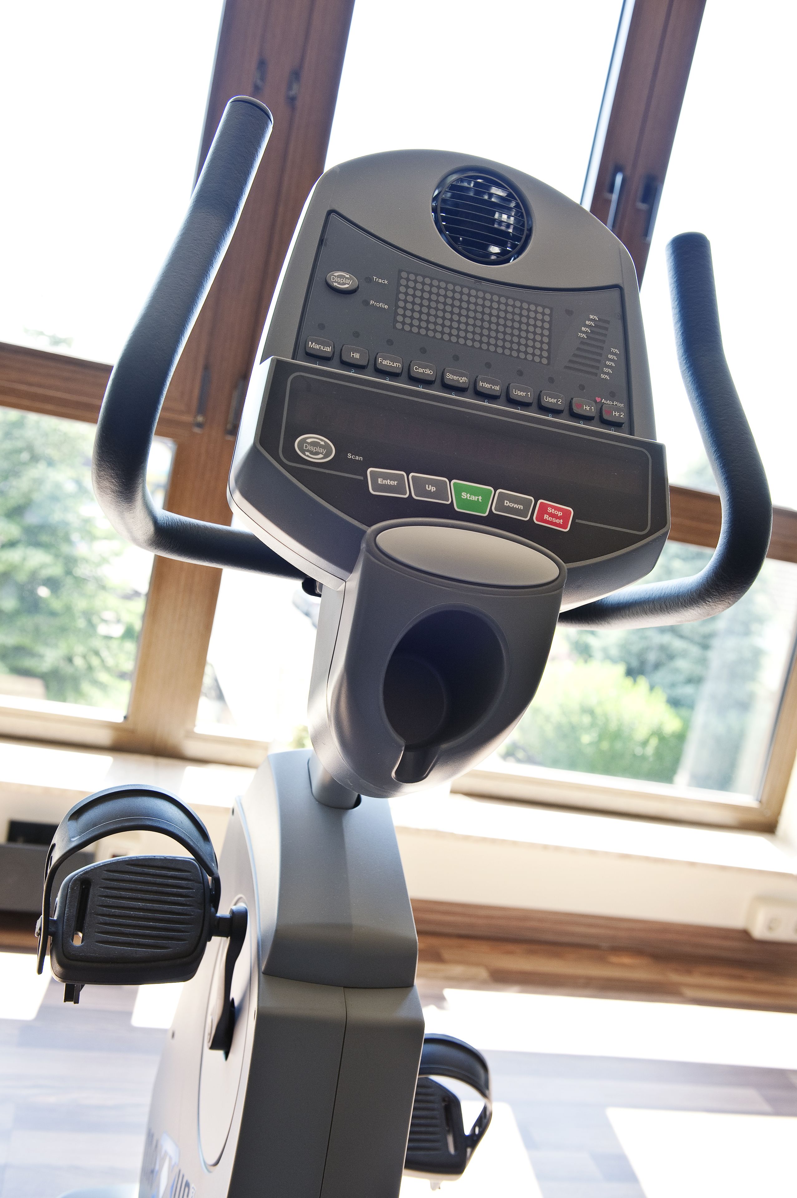 An exercise bike with a digital display and control panel, positioned near a window with a view of greenery outside.