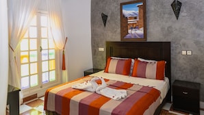 Quarto de hotel com cama, mesa de cabeceira, janela com cortinas, um quadro pendurado na parede e um ornamento decorativo na parede.