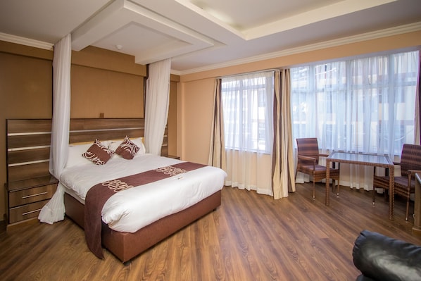Habitación de hotel con una cama grande, suelo de madera, una mesa de comedor con sillas y cortinas translúcidas en las ventanas.