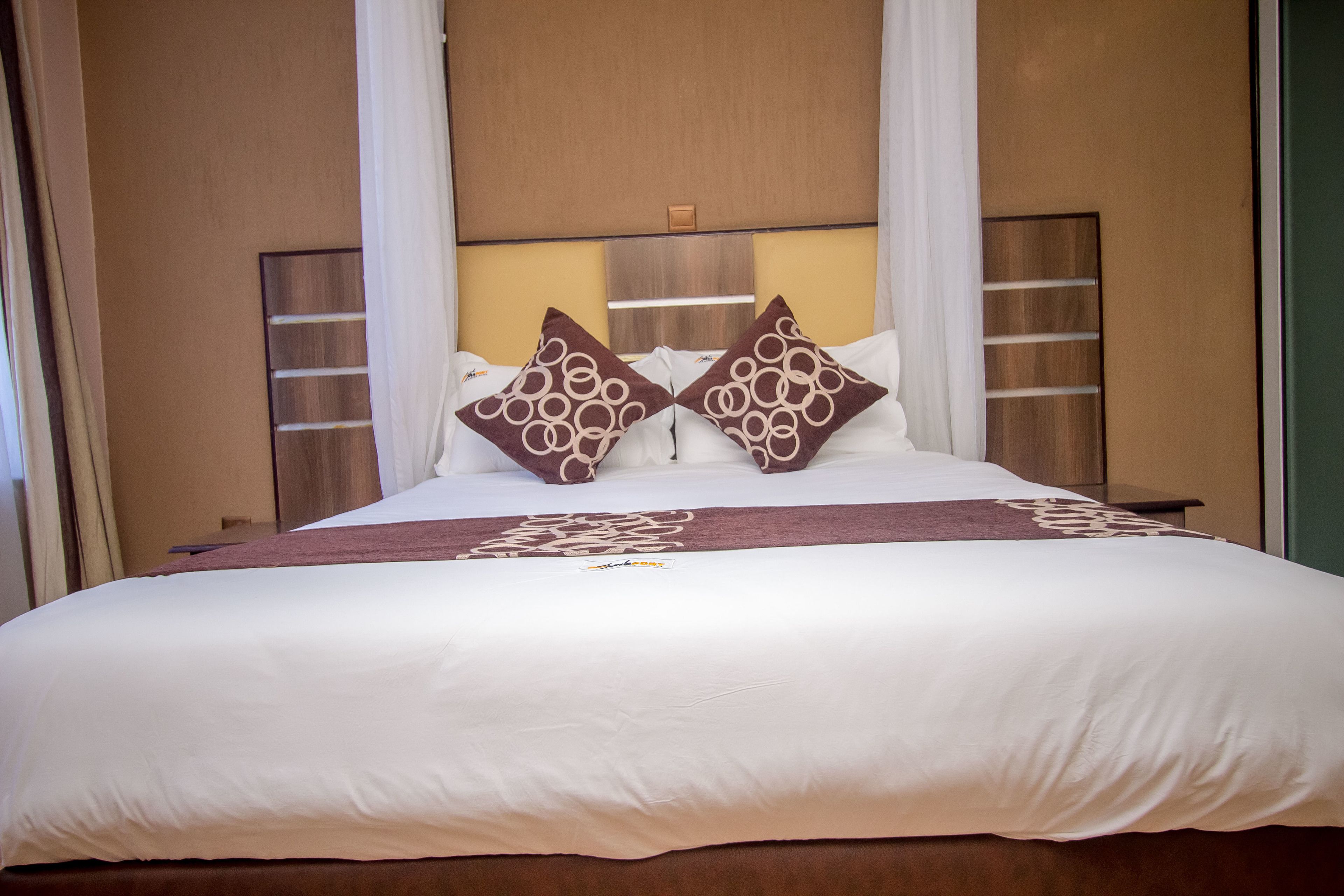 Een net opgemaakt bed met wit en bruin beddengoed, twee decoratieve kussens en een hoofdbord met een lamp.
