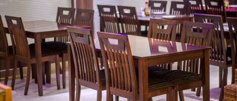 Un comedor con mesas y sillas de madera, piso de baldosas y un ventanal con persianas.