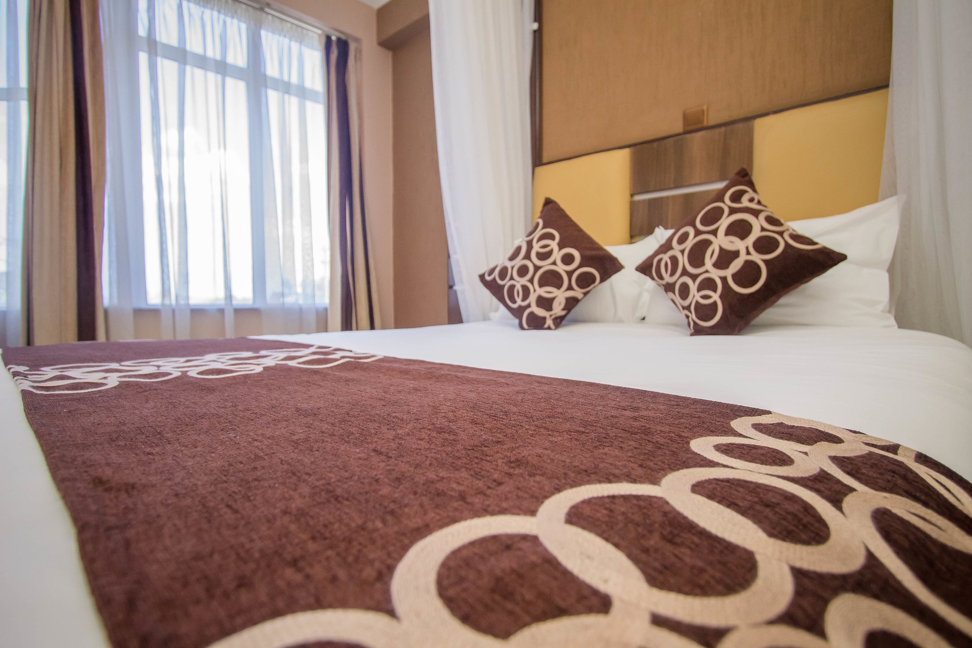 A hotel room with a large bed, two decorative pillows, a window with sheer curtains, and a wooden headboard.