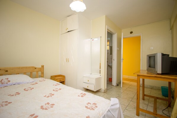 Un dormitorio con cama, armario, televisor sobre una mesa de madera, y un baño visible a través de una puerta abierta.