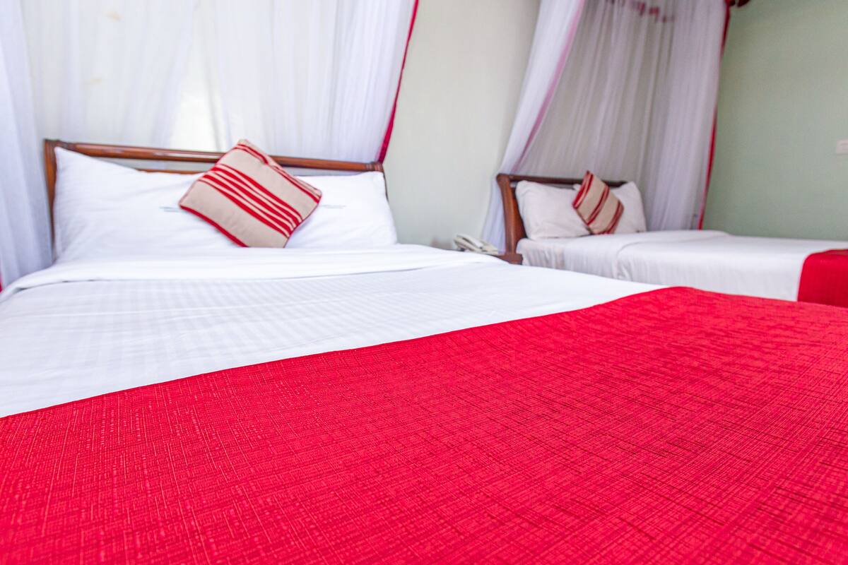 Habitación de hotel con dos camas, cada una con ropa de cama blanca y detalles rojos. La habitación tiene una cama con dosel y otra cama estándar.