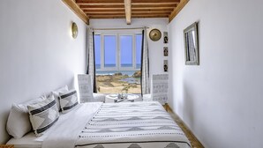 Un dormitorio con cama, paredes blancas, techo de madera, ventana con vista al mar y una mesita con una planta.