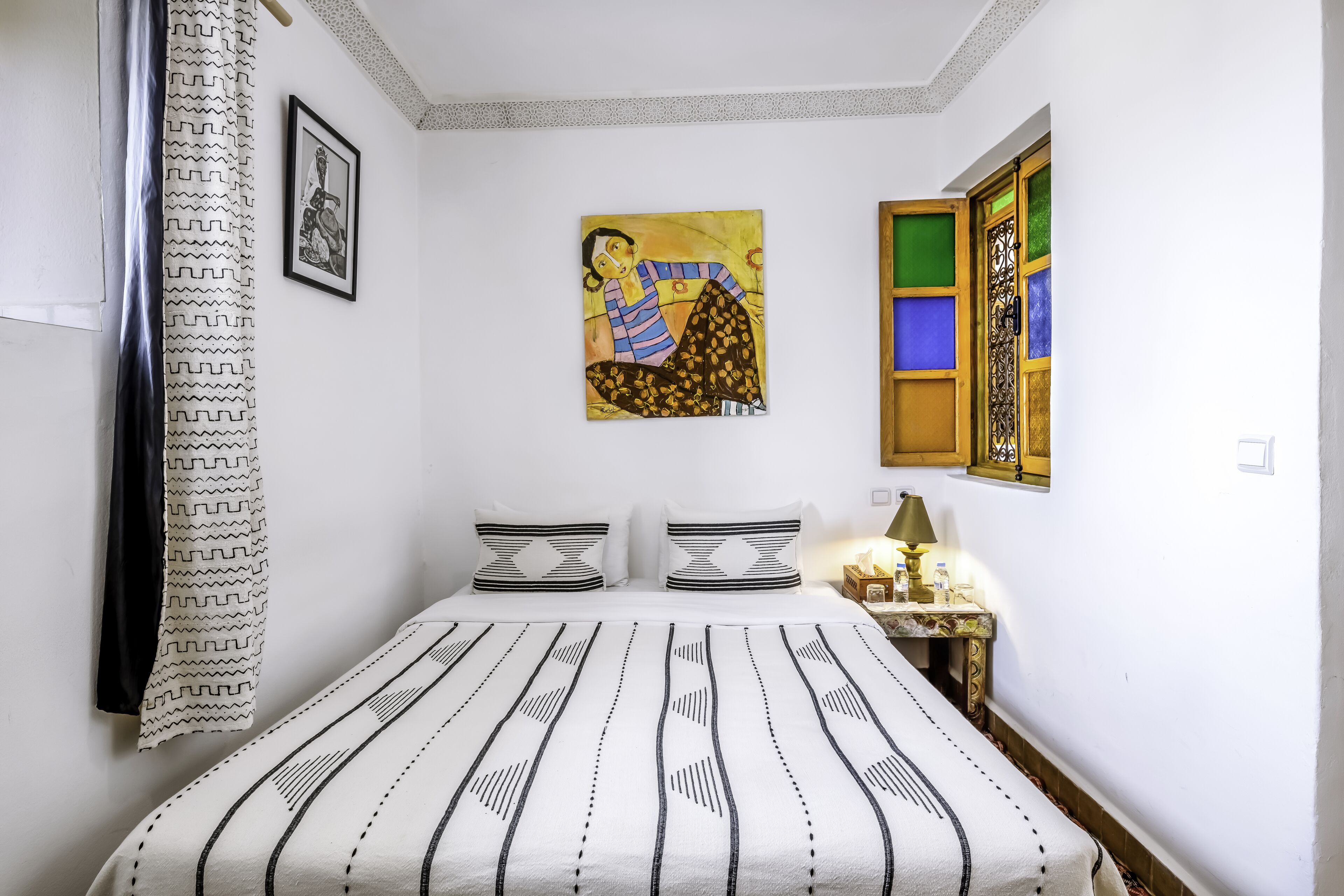 A bedroom with a bed, pillows, a nightstand, a lamp, a painting, and a window with colorful panes.